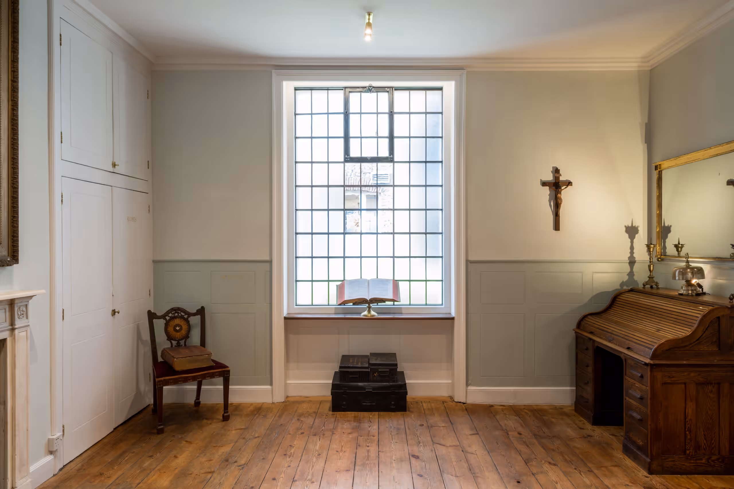 A large window in the centre with an open Bible on the sill. Below the window are three stacked metal boxes. To the right of the window is a desk and a crucifix. To the left of the window is a chair with a large book on the seat.