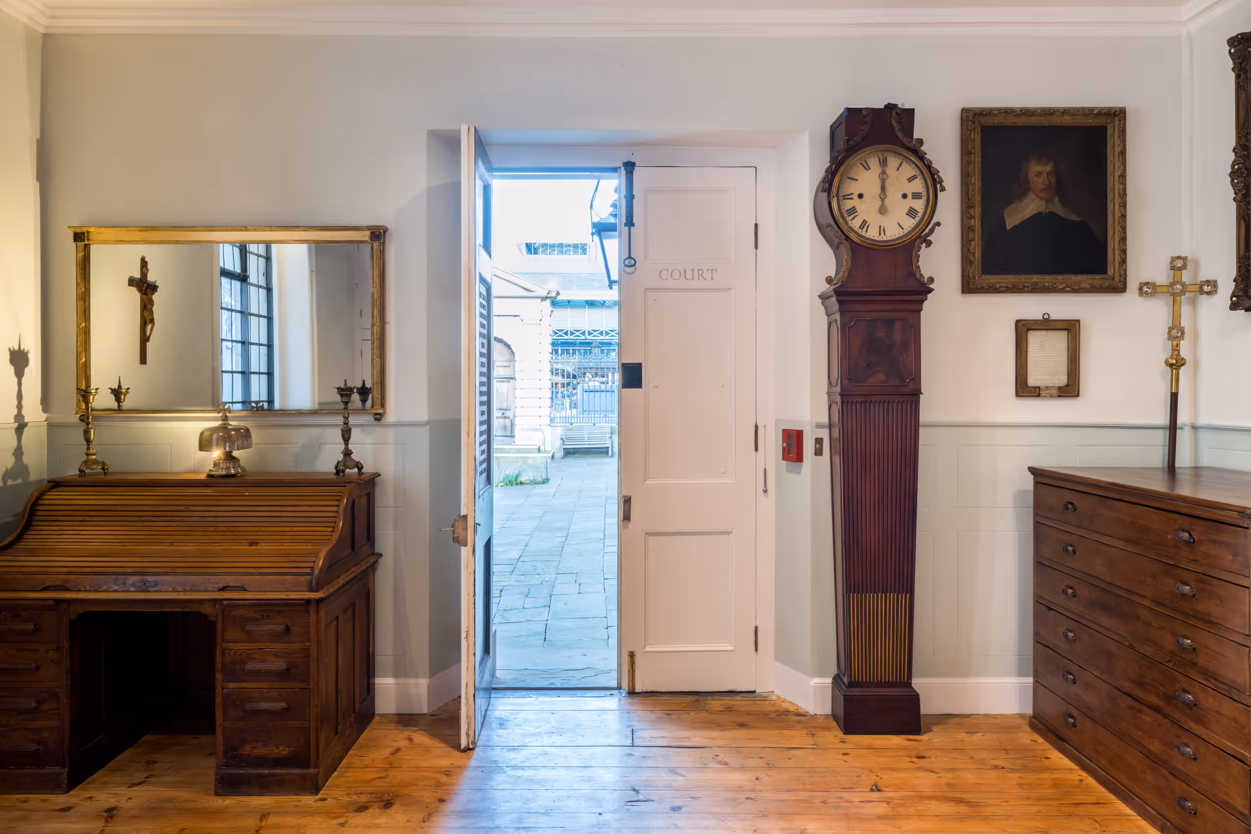 An open door in the centre with a glimpse of the churchyard through it. A large grandfather clock to the right of the door. To the left of the door, a desk with a mirror over it.