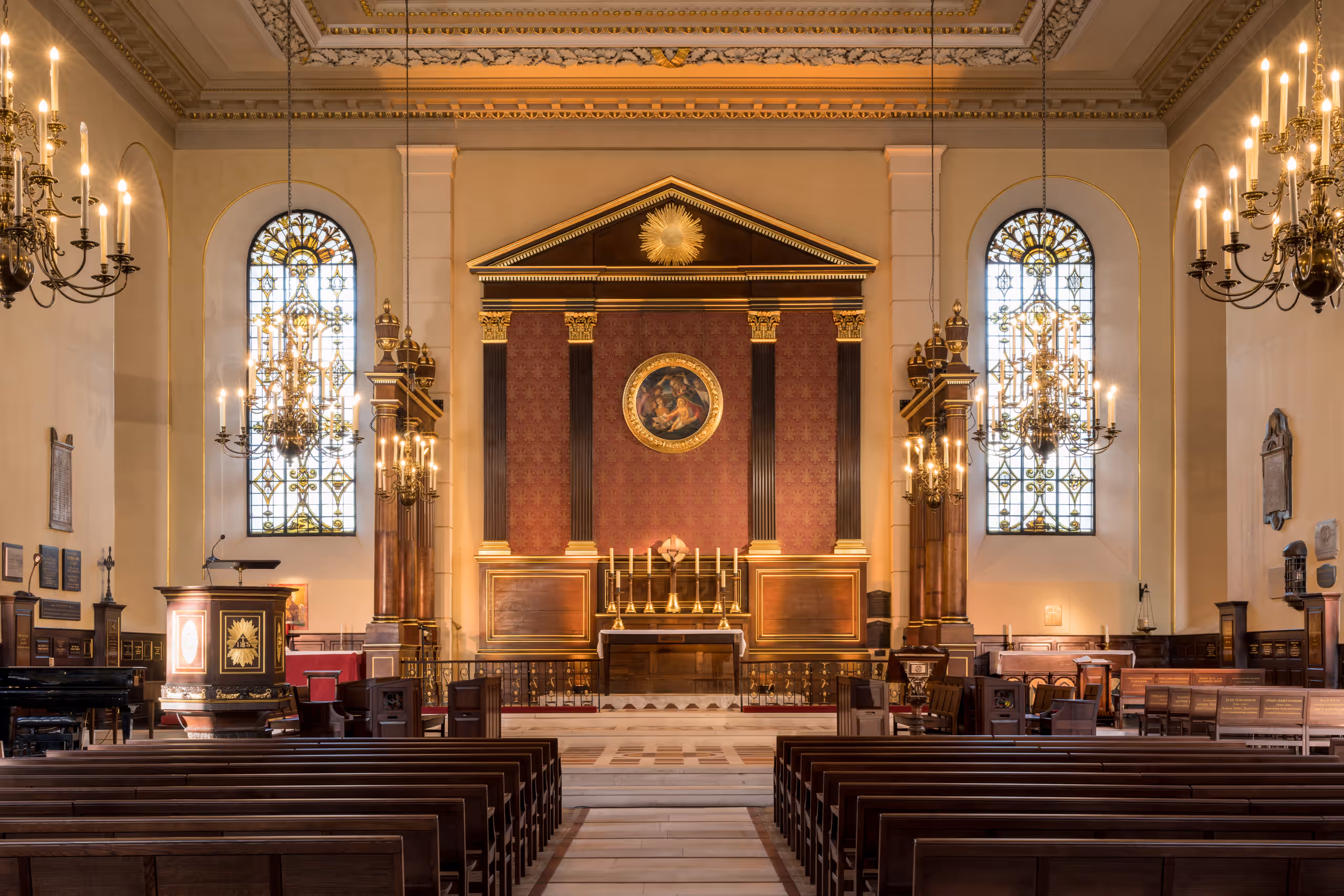 The church interior, with rows of pews leading to the altar between two stained-glass windows and candlelit chandeliers.