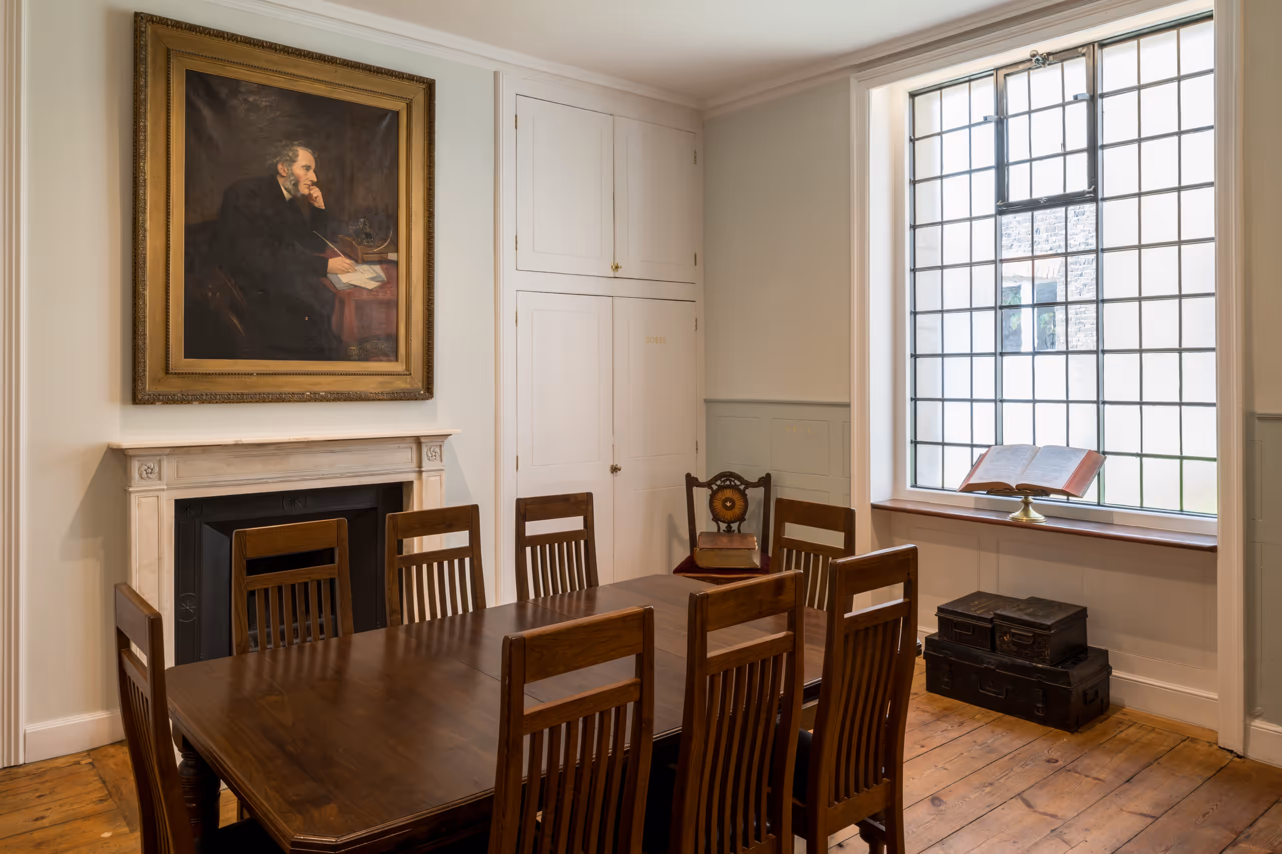 A large wooden table surrounded by chairs in the centre. On the right, a large window with an open Bible on the sill. On the left, a fireplace with a portrait of a Victorian man hung over it.