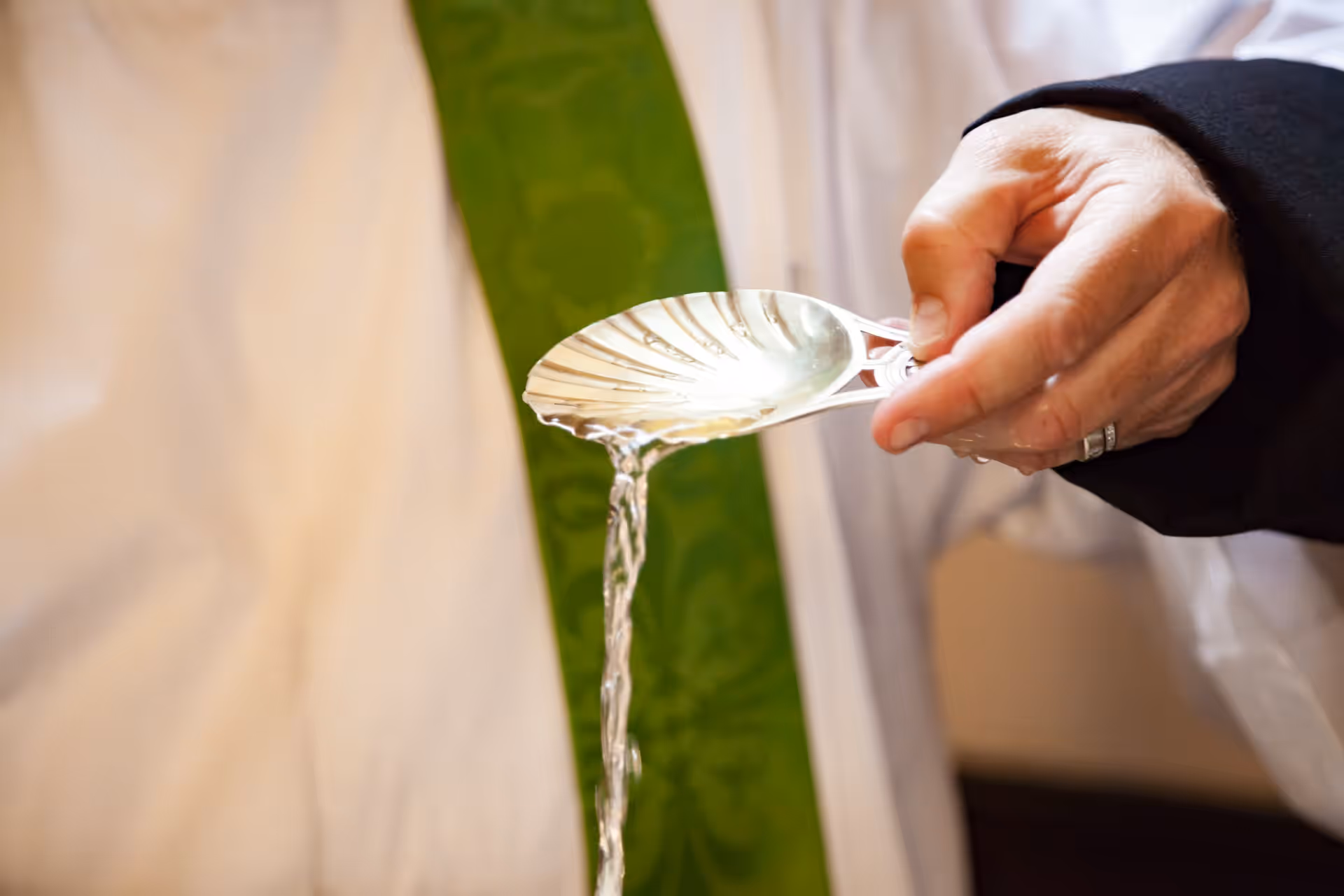Water is poured from a silver shell during a baptism, beside a priest's green stole.