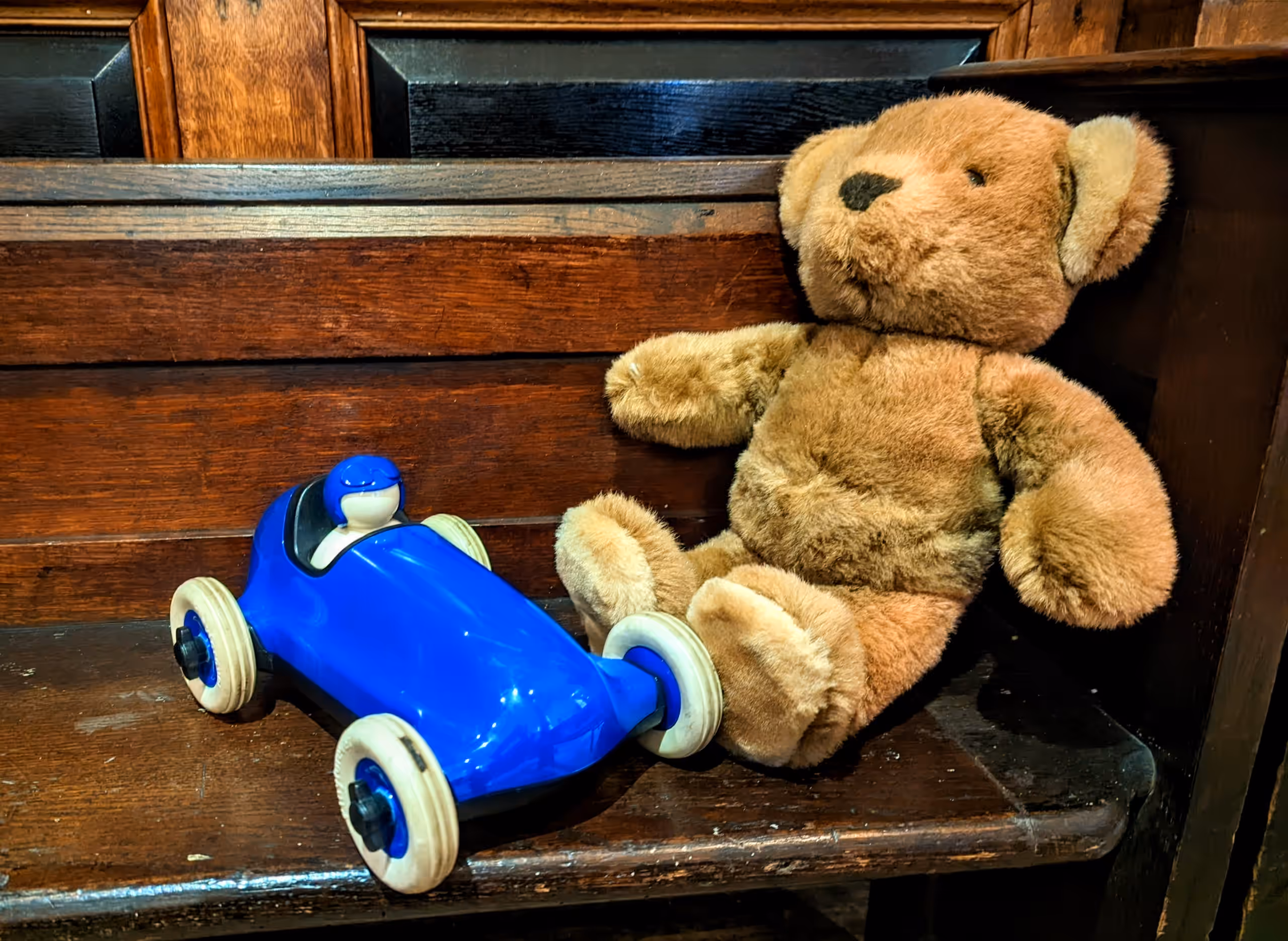 A teddy bear and a blue toy racing car sit side by side on a wooden pew.