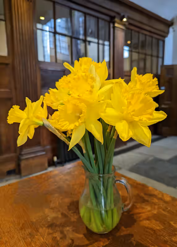 A small glass jug of bright yellow daffodils on a wooden table inside the church.