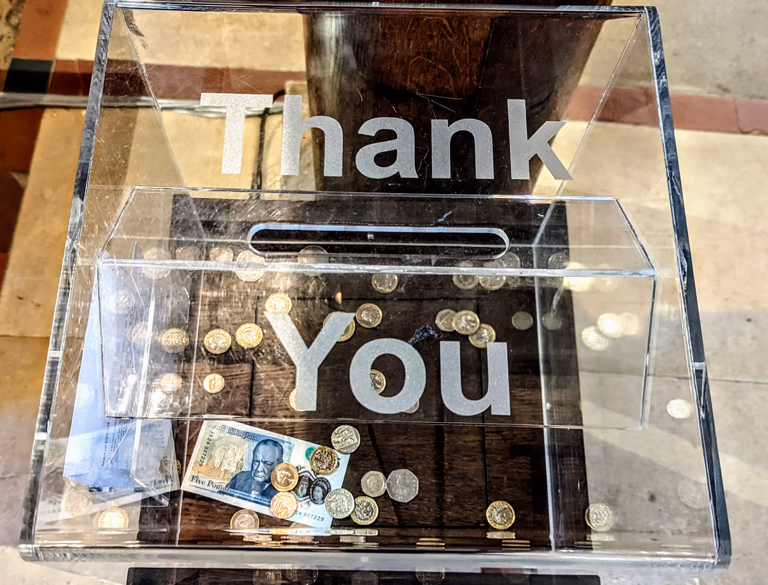 A clear donation box marked 'Thank You', holding coins and a banknote.