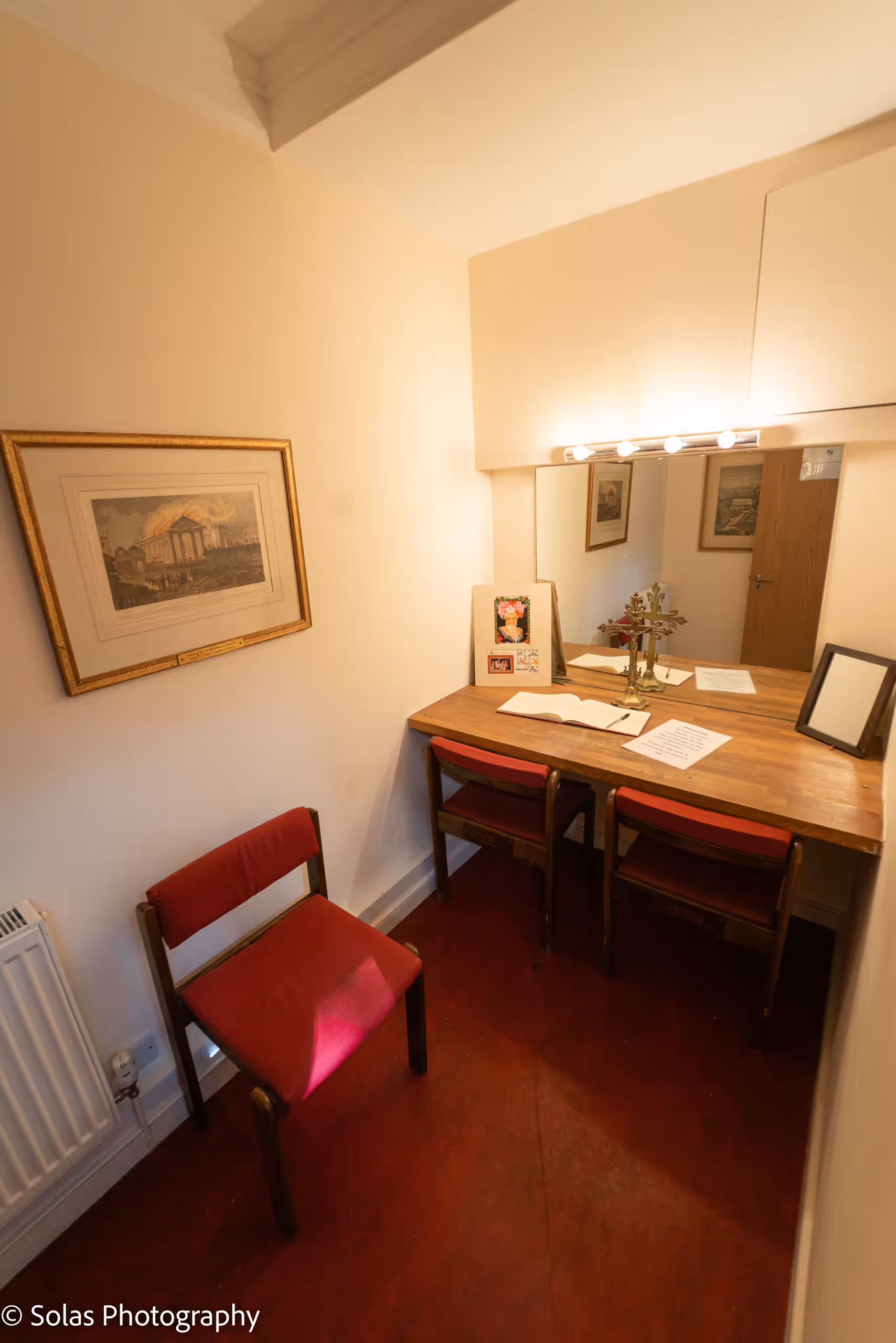 A small dressing room with a mirrored dressing table under a strip of bulbs, red chairs and a framed print.