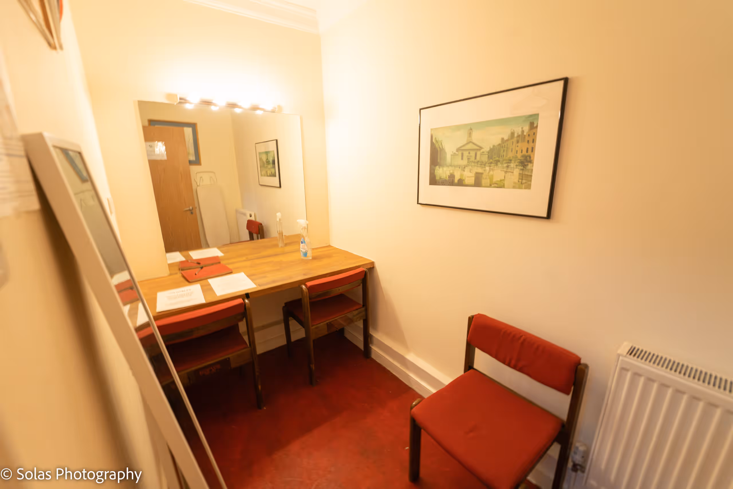 A dressing room with a lit mirror above a shared dressing table, red chairs and a framed print of the church.