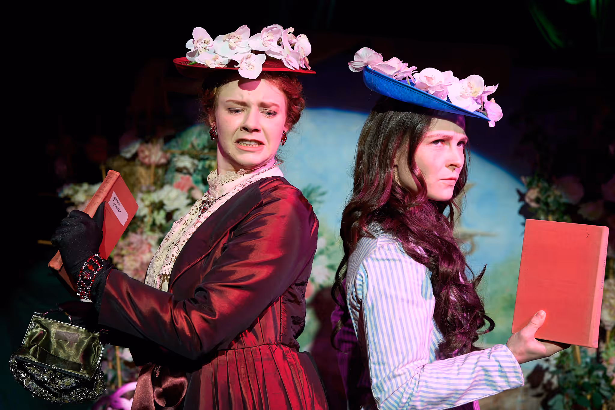Two actresses in Victorian costume and flowered hats stand back to back, each holding a red book.