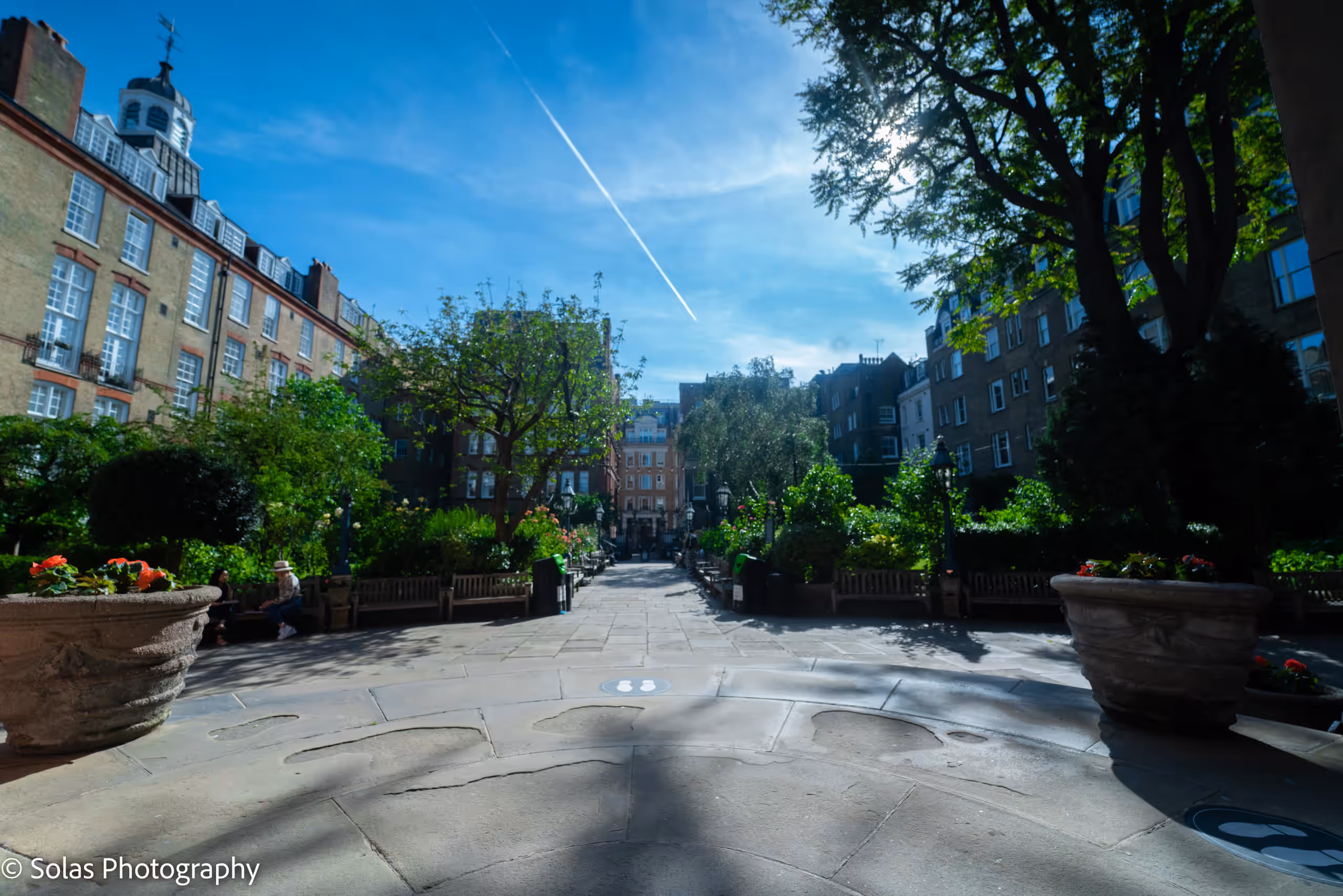 A wide paved path runs through the church garden between planters, benches and trees, with the surrounding buildings beyond.