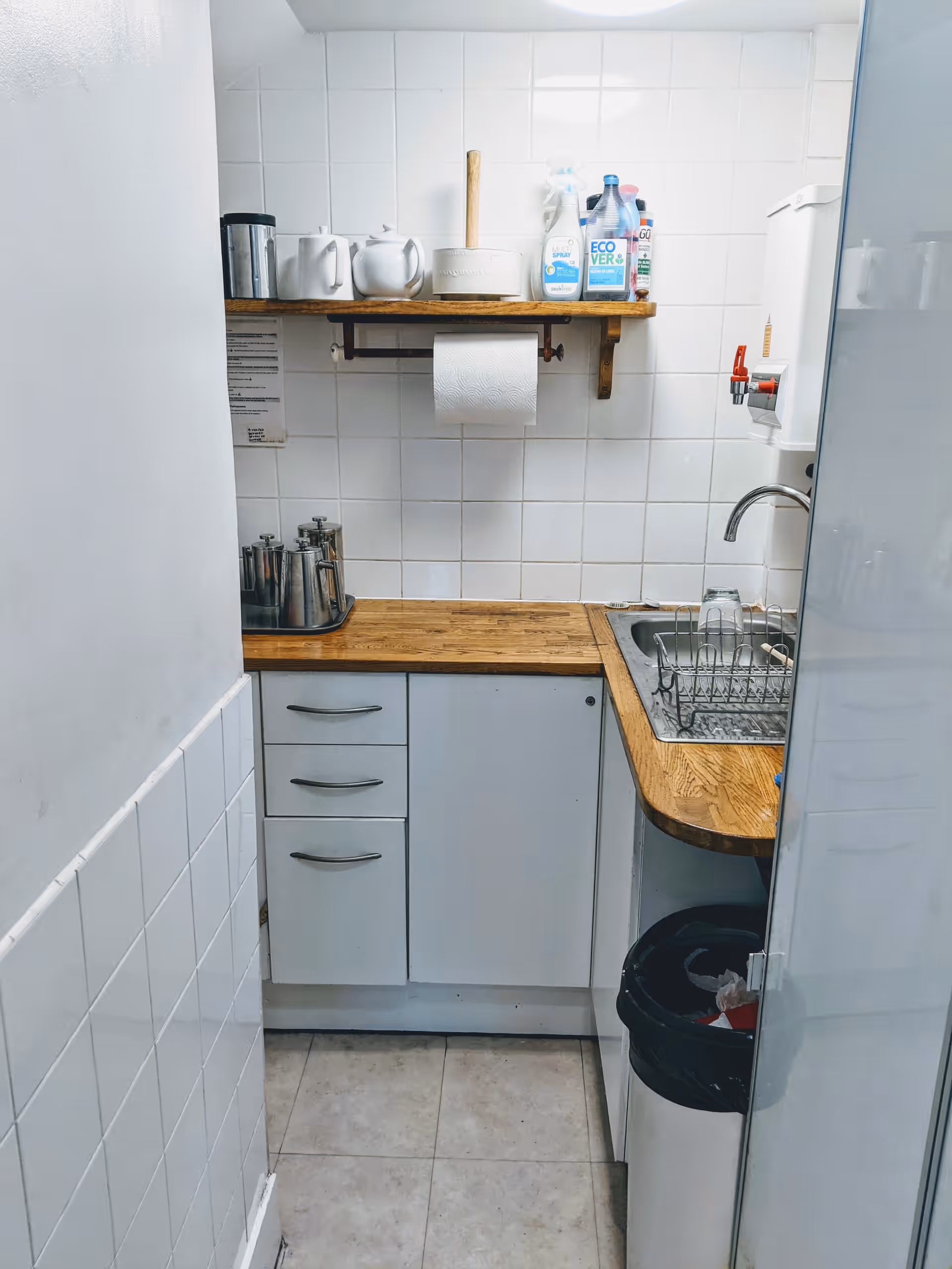 Kitchen cupboards and surfaces. A sink to the right and a shelf above with kitchen supplies.