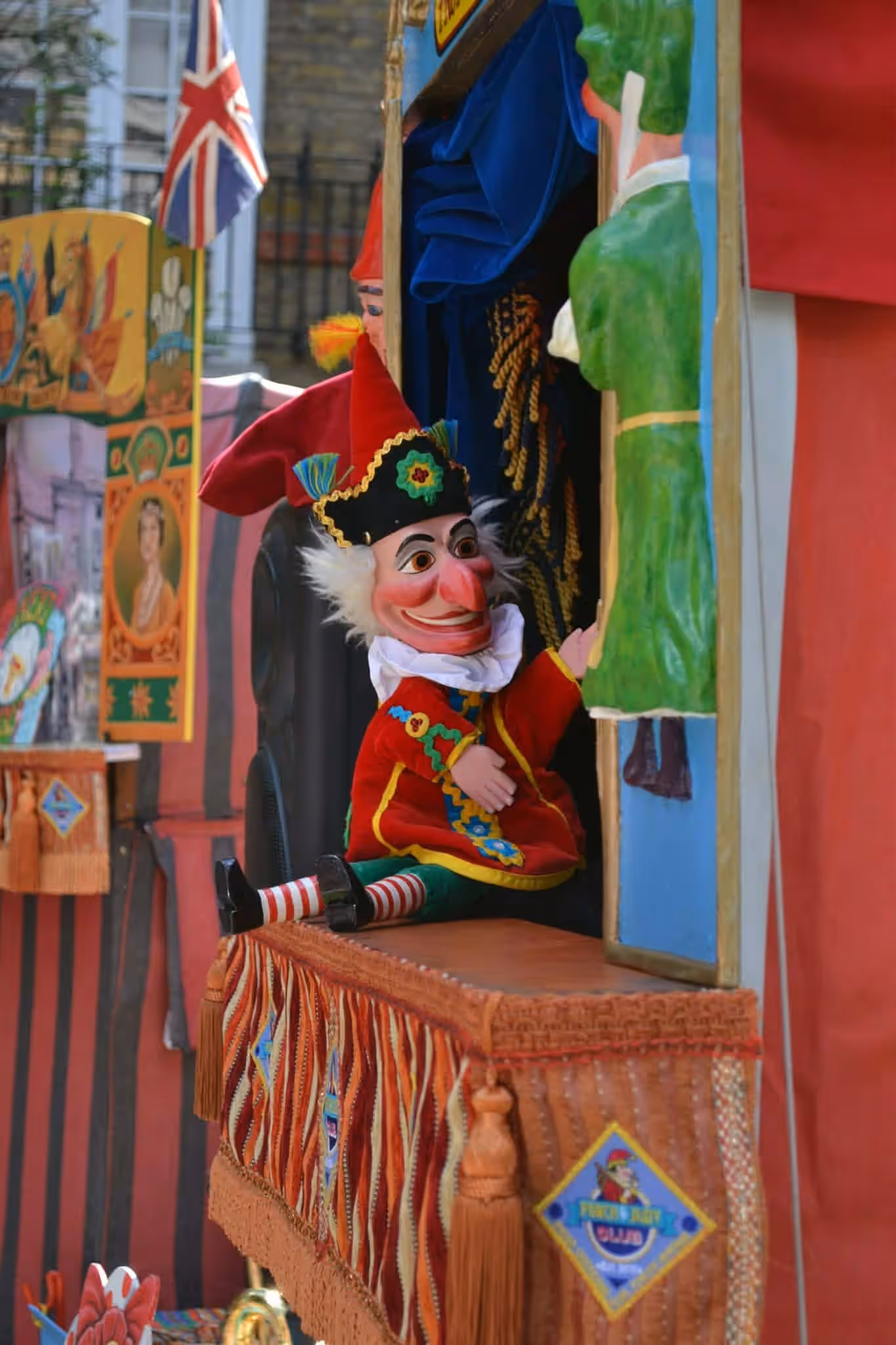 A Mr Punch puppet in a red and gold costume leans out of a puppet booth at the May Fayre.