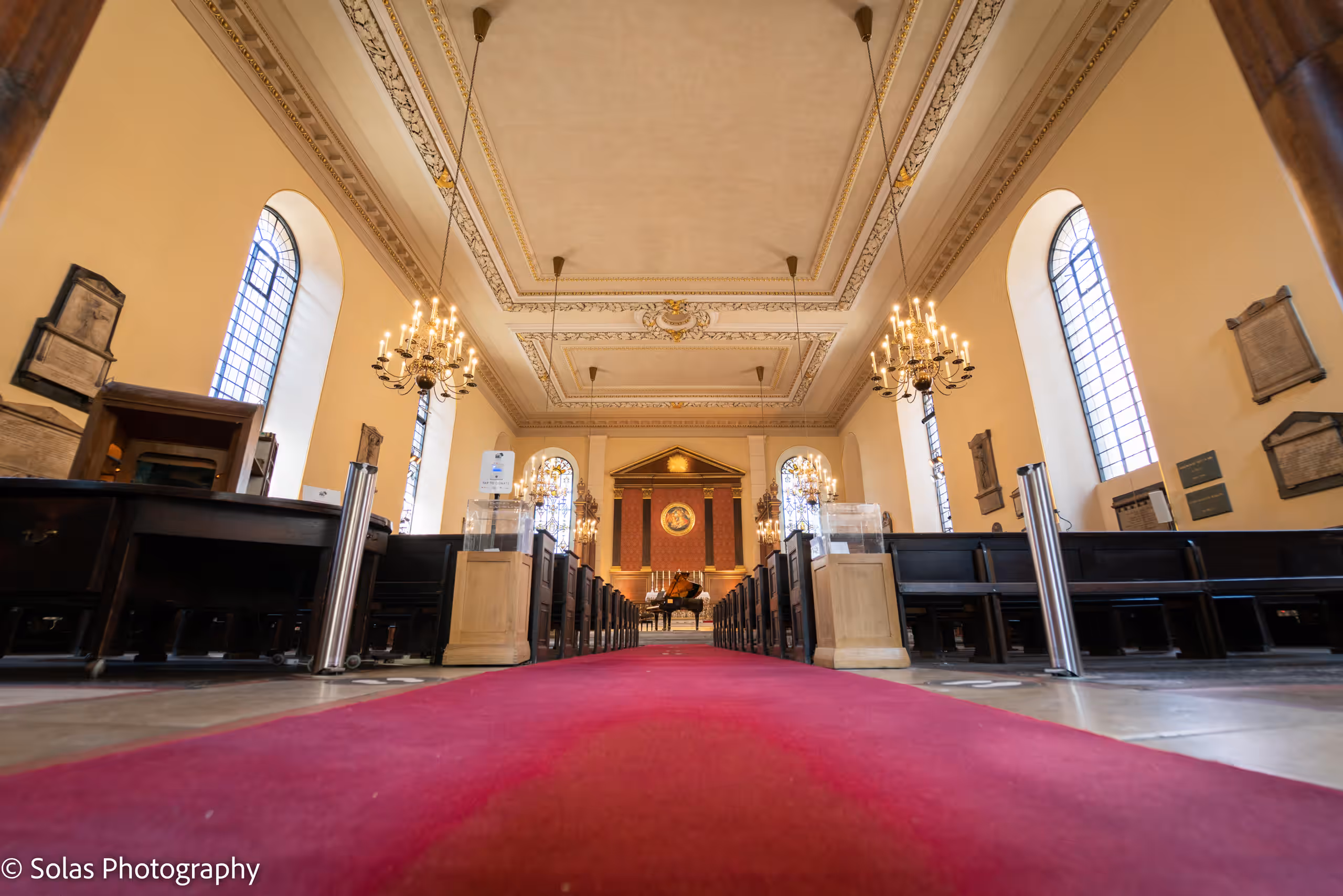 The church interior from the aisle, a red carpet leading between pews and chandeliers towards the altar.