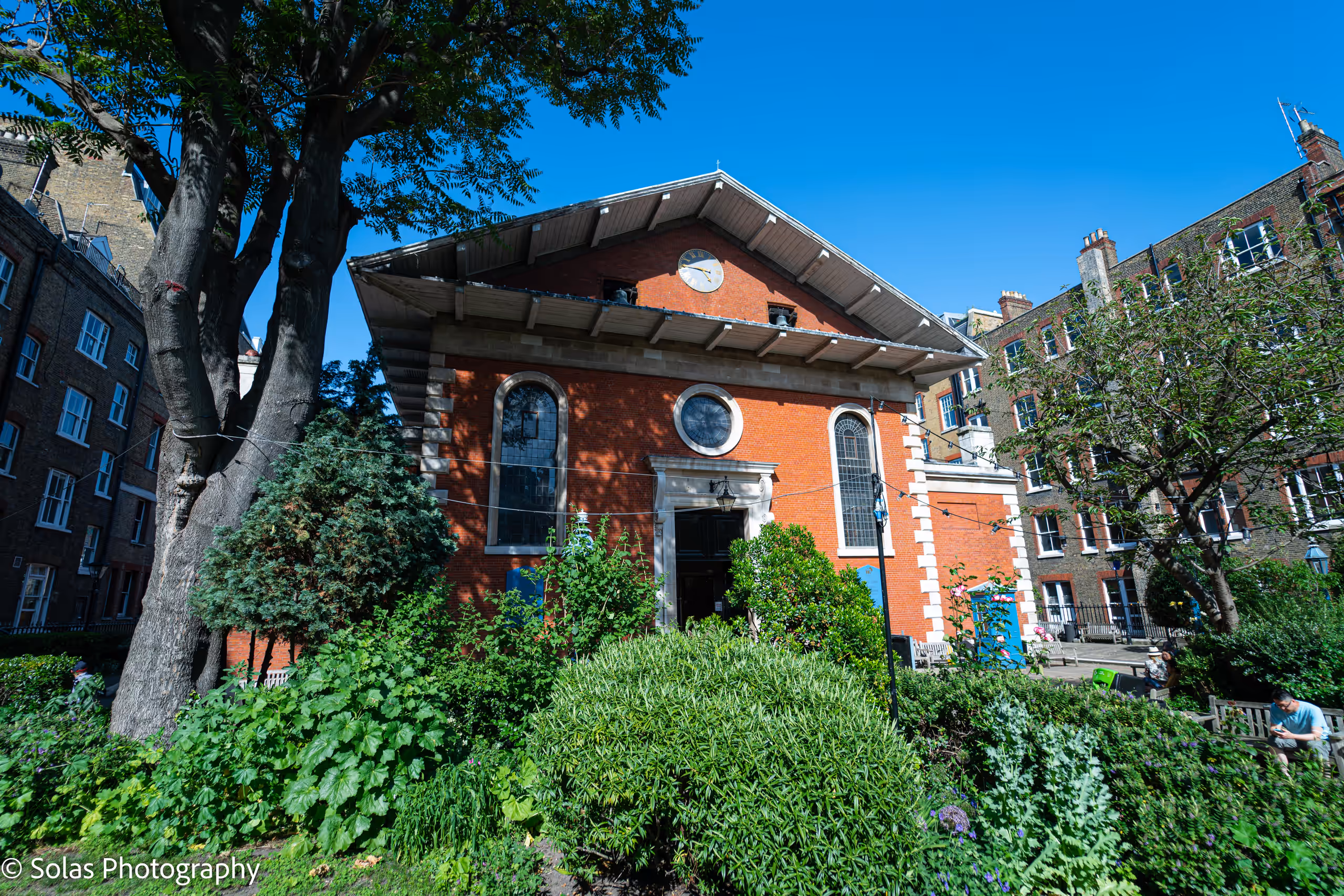 The church's red-brick end wall, with its clock and arched windows, seen across the leafy church garden on a sunny day.