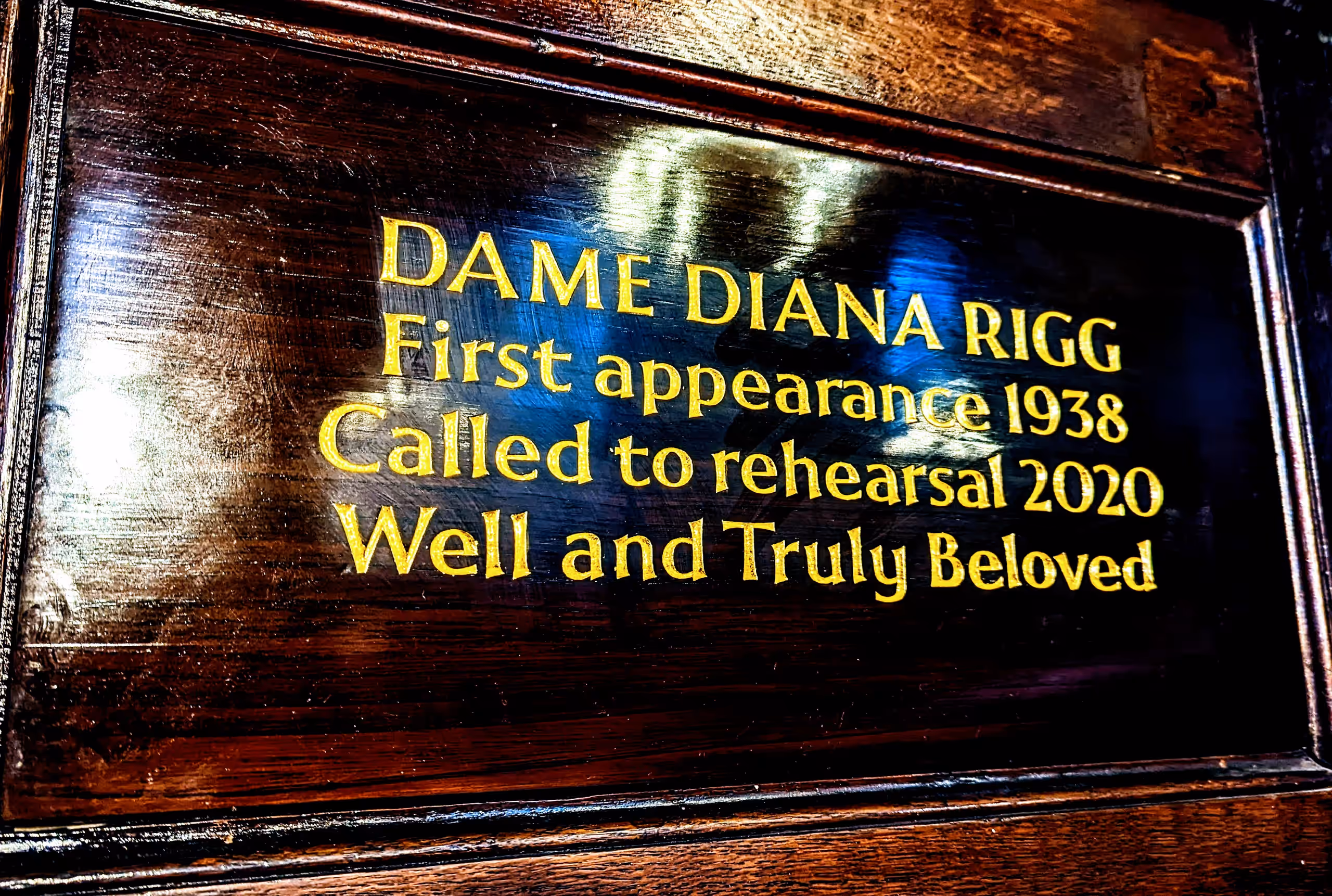 Gold lettering on a dark wooden memorial plaque: 'Dame Diana Rigg. First appearance 1938. Called to rehearsal 2020. Well and Truly Beloved.'