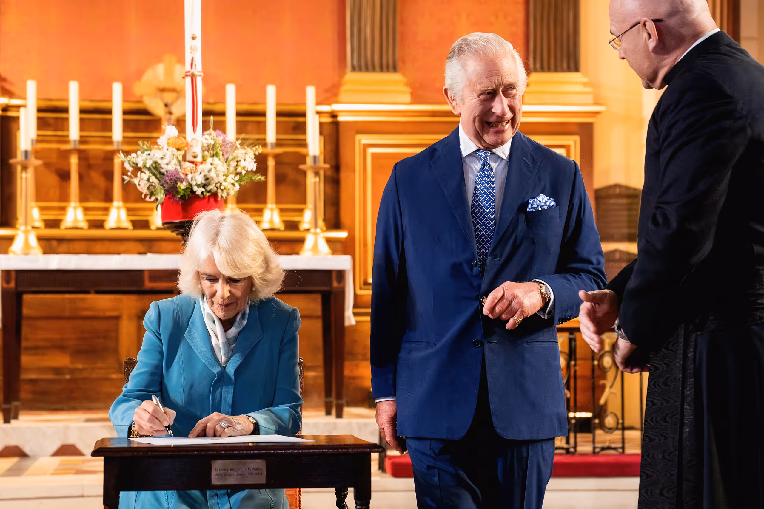 Queen Camilla signs a commemorative page at a table before the altar as King Charles laughs beside her.