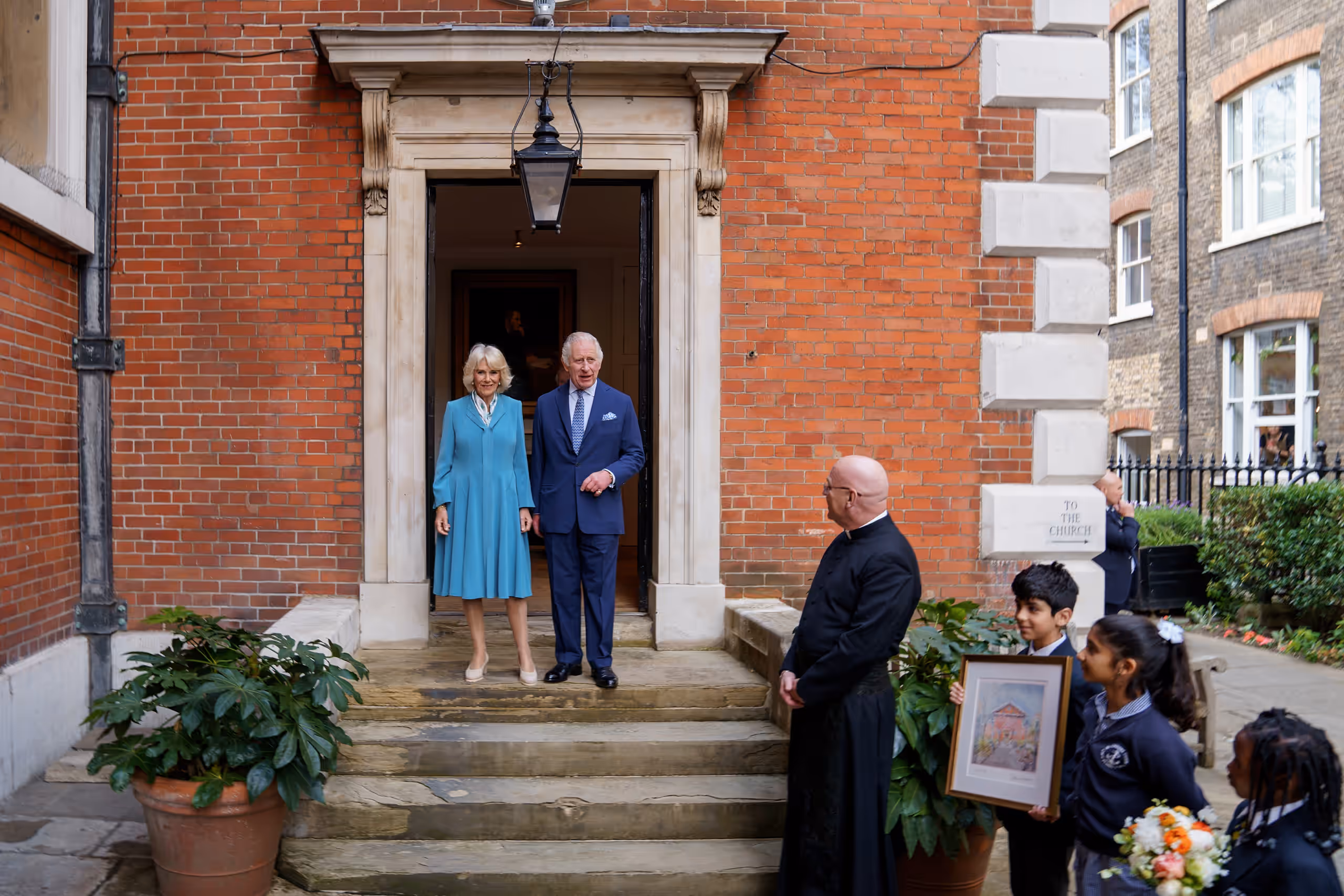 The King and Queen at the vestry door as schoolchildren wait with flowers and a framed painting of St Paul's.