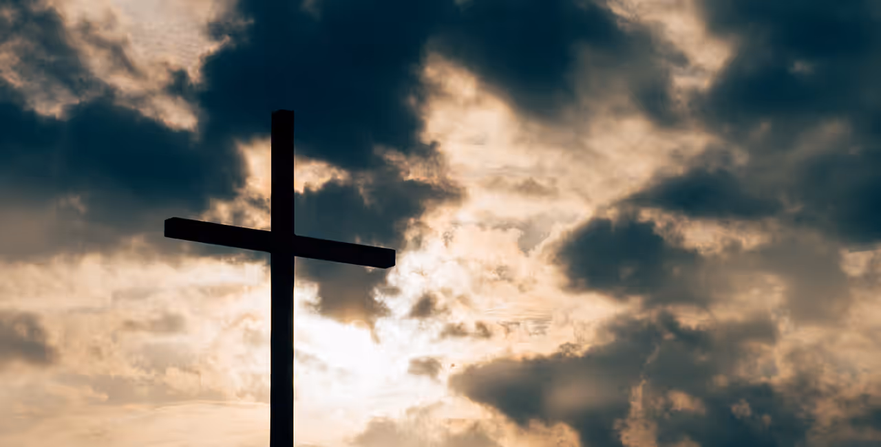 A wooden cross silhouetted against a cloudy sky with sunlight breaking through.